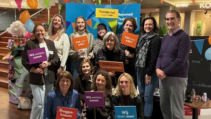 groepsfoto met feestelijke vlaggen en de mensen houden bordjes omhoog met teksten als "Wij werken samen.", "Wij doen mee", en "Tijd voor actie" .